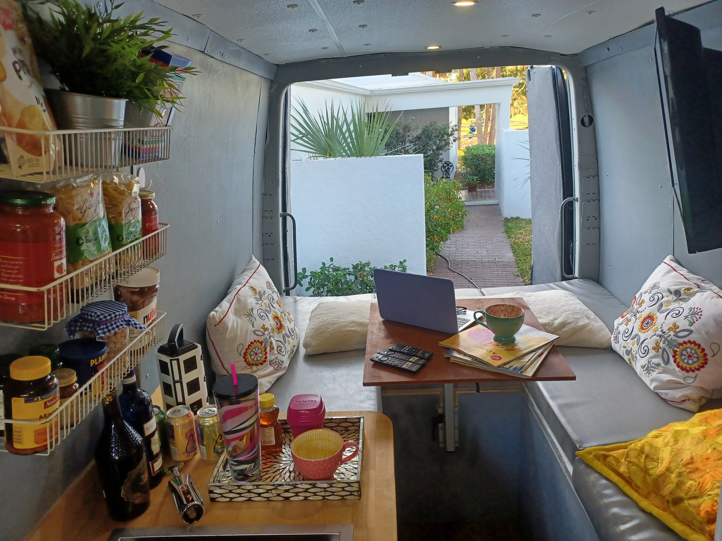 Cozy rv rental interior featuring a compact kitchen, bed, and laptop workspace. Open doors reveal a serene outdoor view.