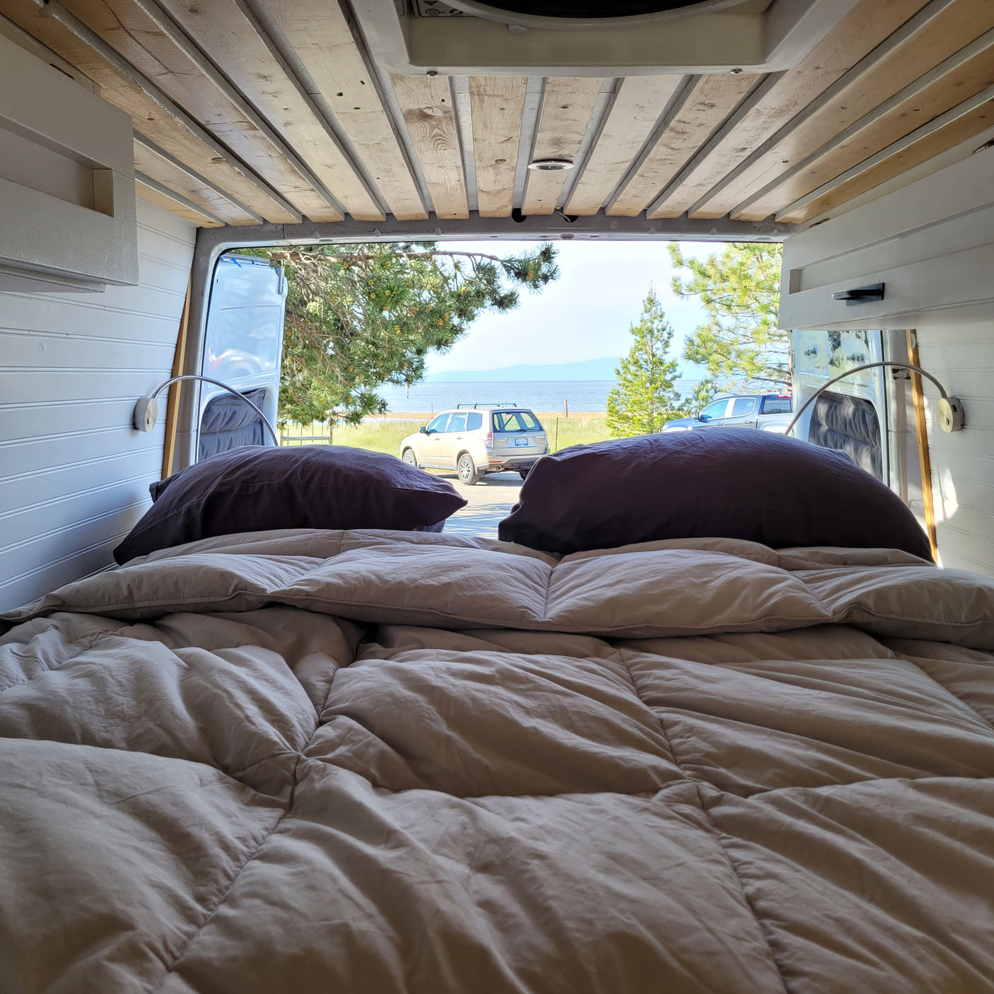 Experience a serene rv rental. View from a cozy van bed shows a lake, mountains, and parked vehicles under blue sky.