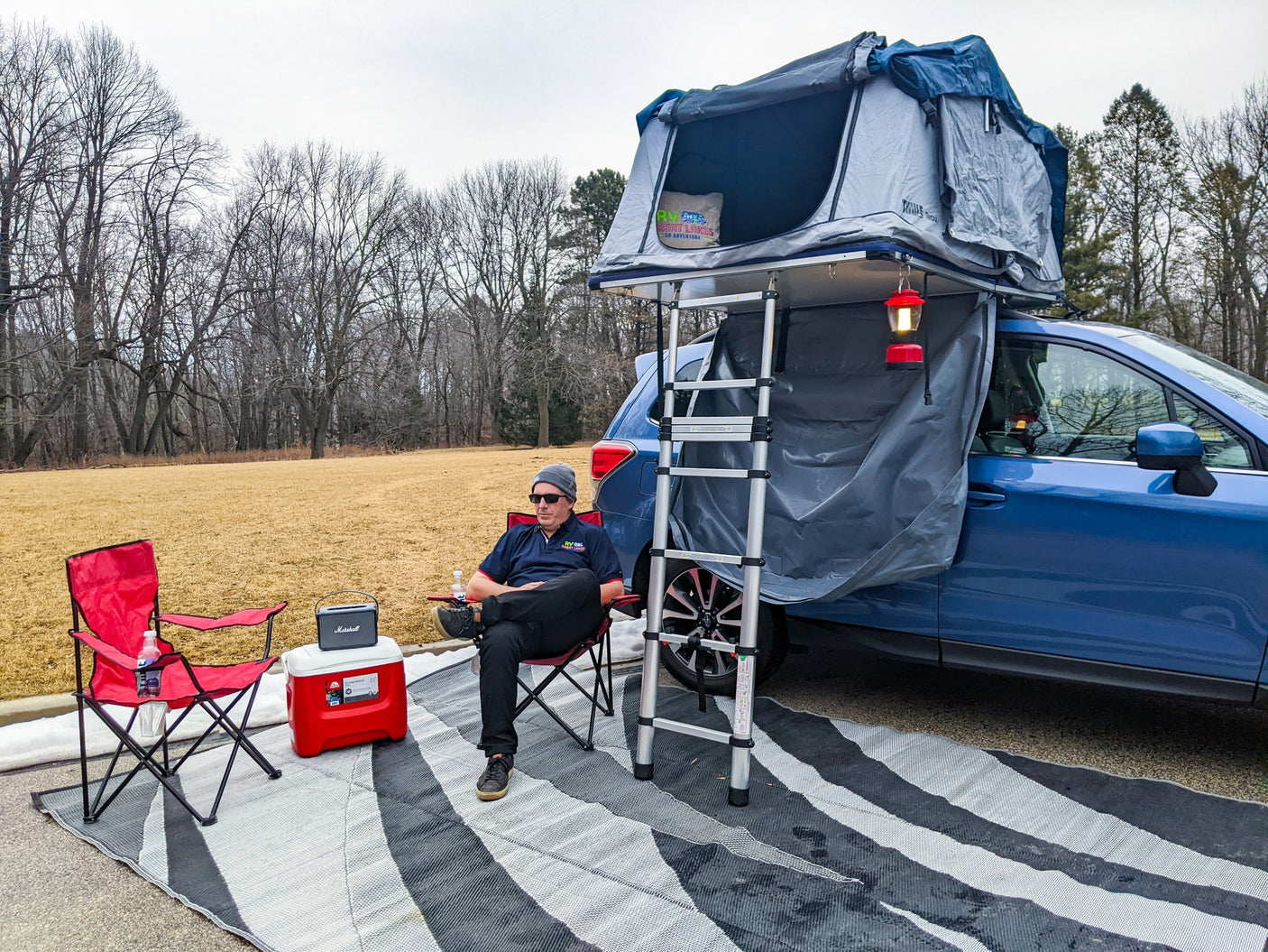 Perfect for an RV rental getaway! A man relaxes beside his car-top tent setup with cooler, speaker, and camping gear.