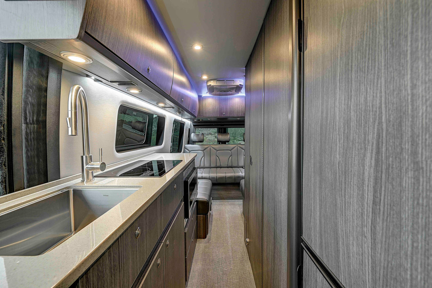 Modern RV rental interior: a sleek kitchen with a sink, induction cooktop, cabinets, and a cozy rear lounge, well-lit.