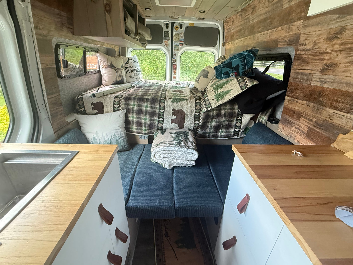 Cozy RV rental campervan interior featuring a comfy bed with nature-themed decor, kitchen, and wood paneling.