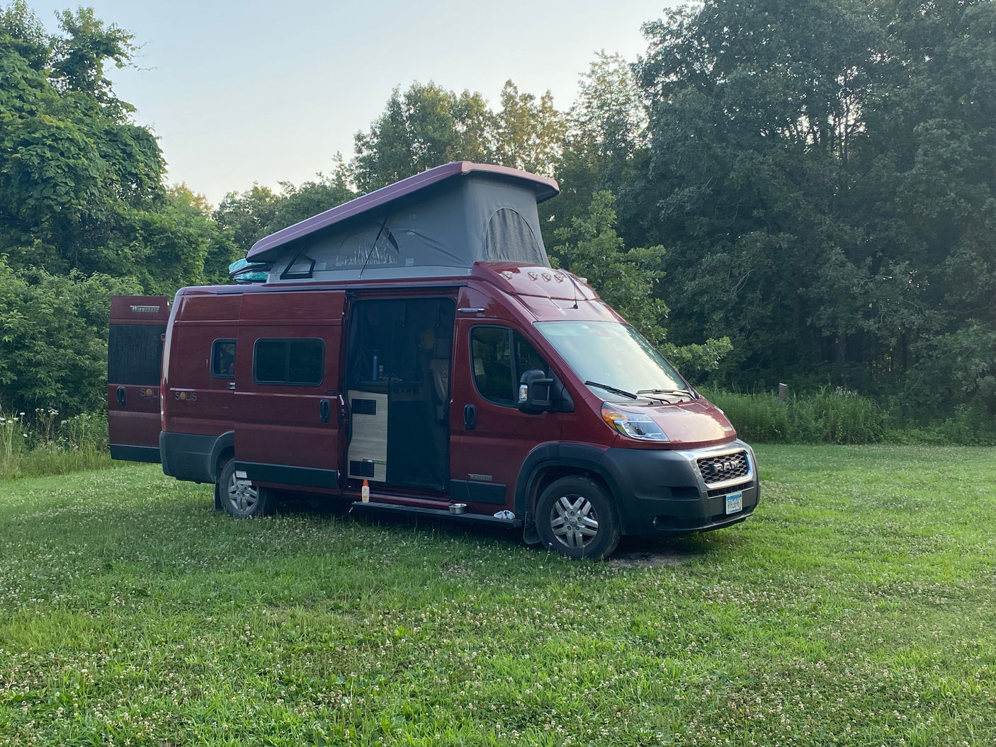 A red Winnebago Solis camper van with an extended pop-top is parked in a grassy field, perfect for an RV rental outdoor adventure.