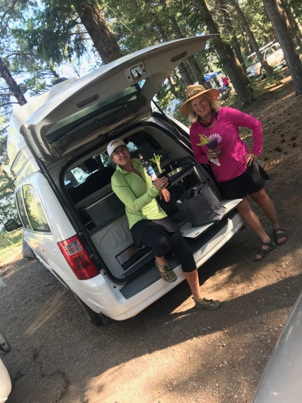 Two women smile by their open RV rental, showcasing a comfortable camp setup in a sunny forest. Ready for adventure!