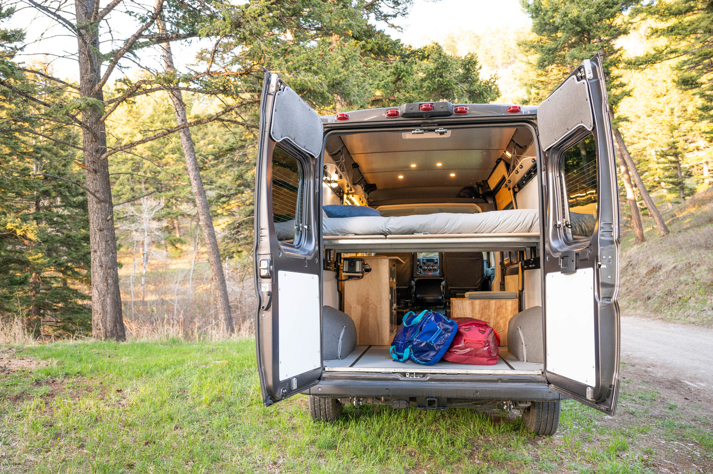 Open rv rental camper van rear view. Shows cozy bed, storage with bags, and illuminated interior, parked in a forest.
