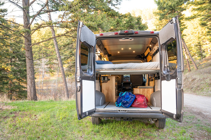 Open rv rental camper van rear view. Shows cozy bed, storage with bags, and illuminated interior, parked in a forest.