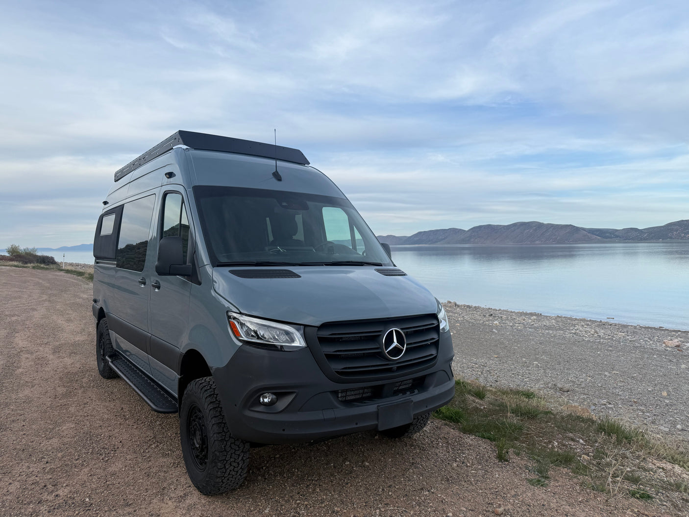 Experience freedom! Grey Mercedes-Benz Sprinter camper van, perfect RV rental, parked by a scenic lake with distant mountains.