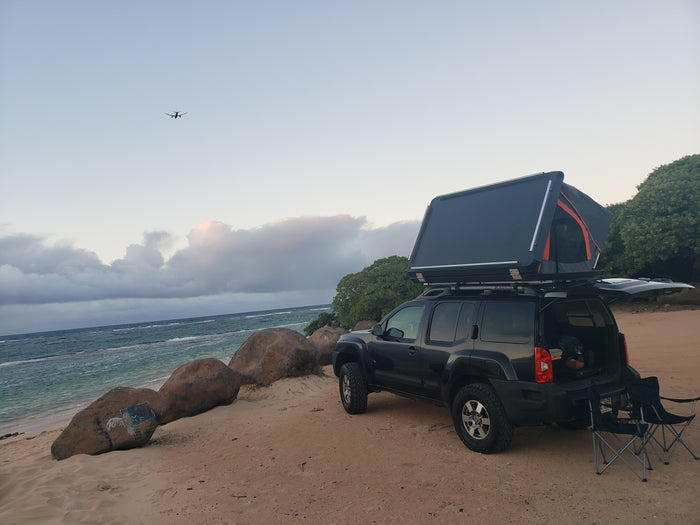 RV rental style: SUV with rooftop tent for beach camping. Chairs by the ocean, plane overhead.