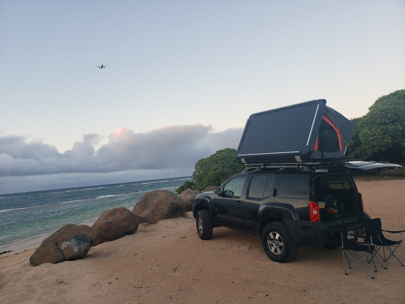 RV rental style: SUV with rooftop tent for beach camping. Chairs by the ocean, plane overhead.