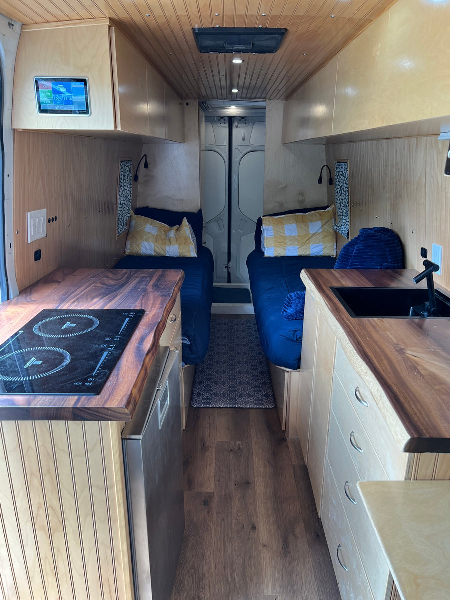 This RV rental interior features a compact kitchen with cooktop/sink, cozy twin beds, and smart controls, perfect for travel.
