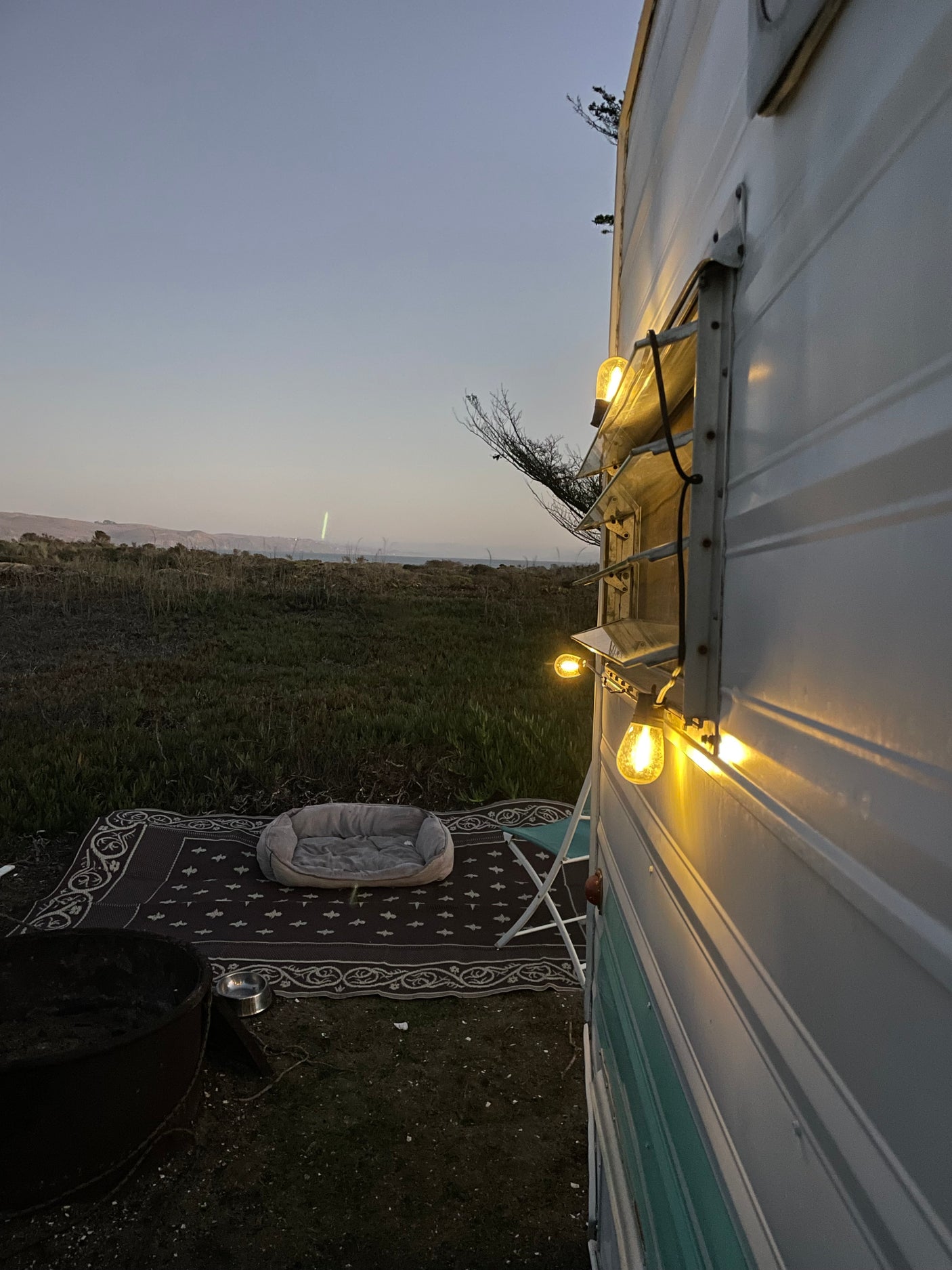 Enjoy a relaxing evening at your RV rental campsite. String lights illuminate a pet bed and fire pit under the twilight sky.