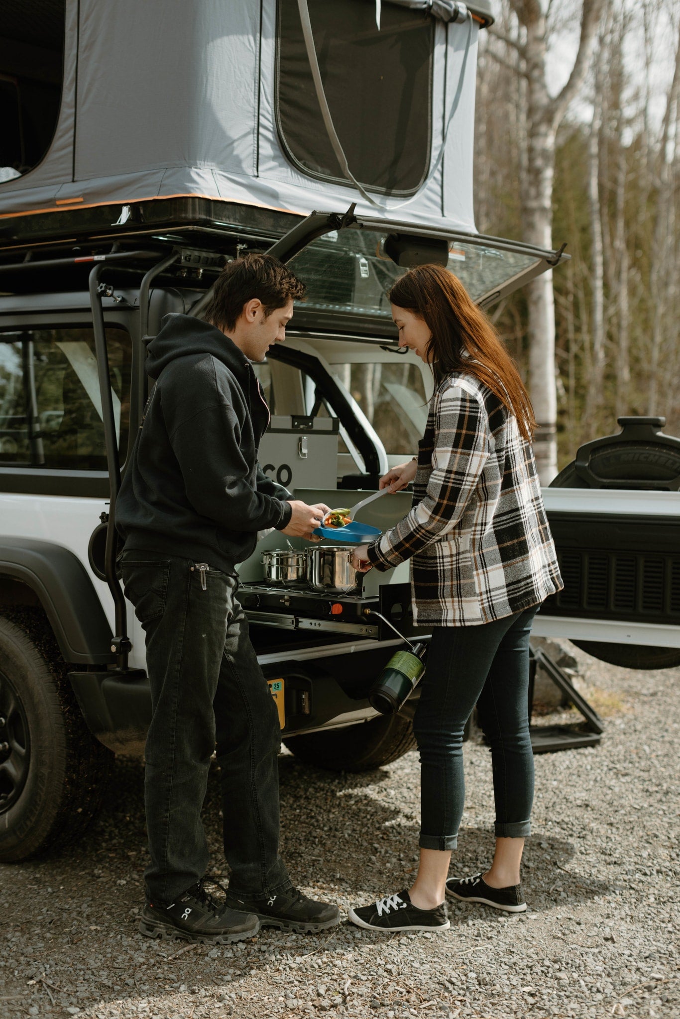 A couple cooks a meal using an RV rental's pull-out camp kitchen, attached to a white vehicle with a rooftop tent.