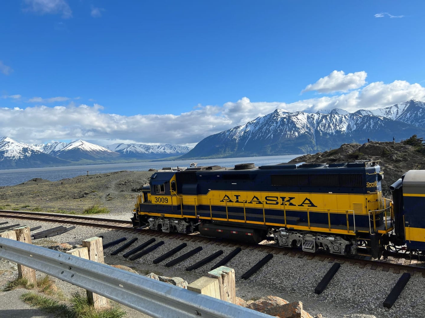 Alaska's majestic scenery with a blue and yellow train (3009) by snowy mountains & water. Explore by train or find an RV rental!