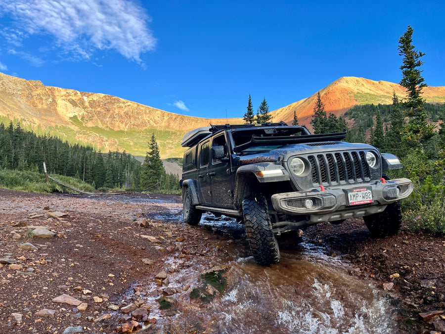 A black Jeep Gladiator with rooftop tent tackles a rocky mountain trail and stream. An epic scene for your next RV rental adventure!