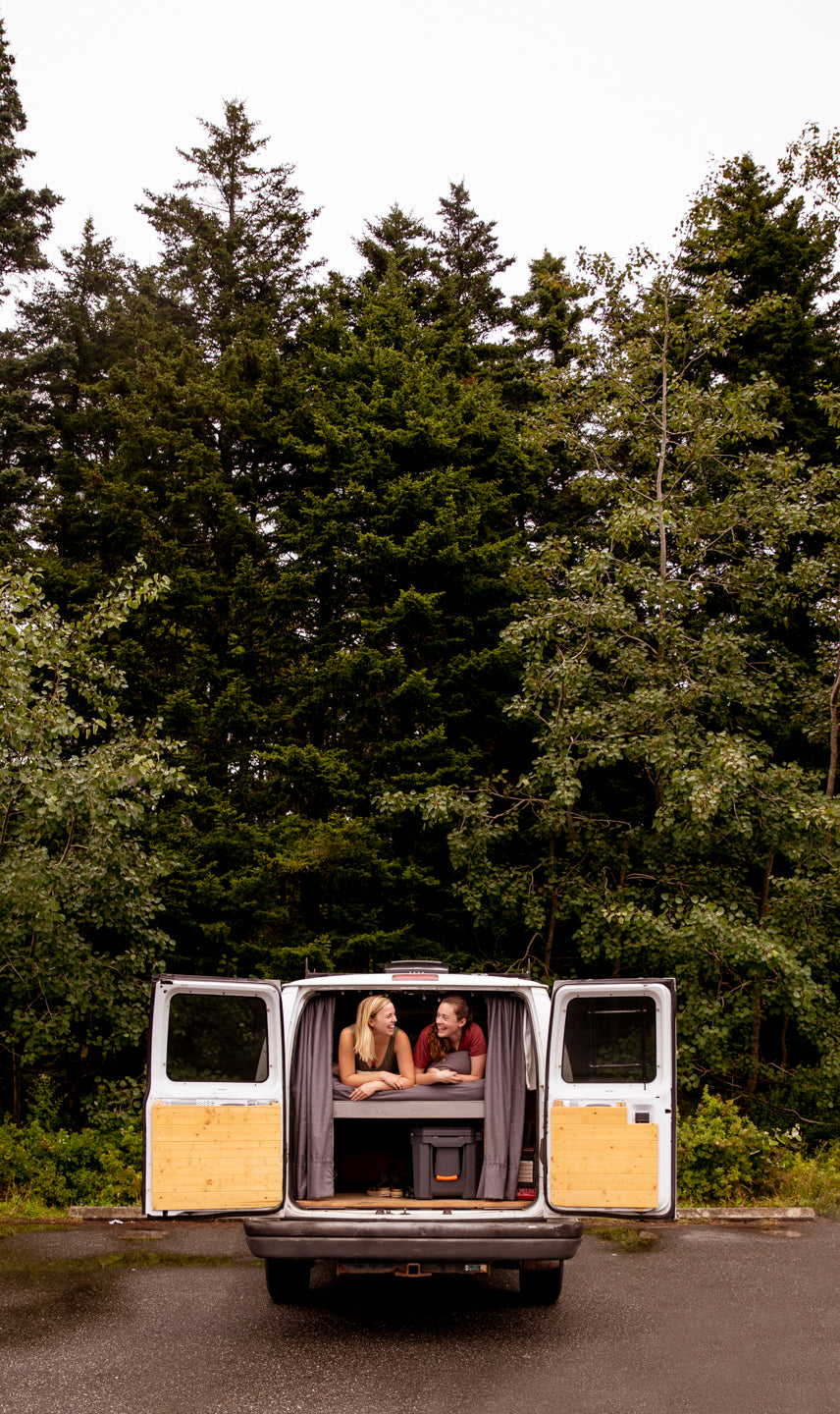 Two women laugh in their open camper van, parked on wet ground in a forest. Perfect RV rental for road trips!