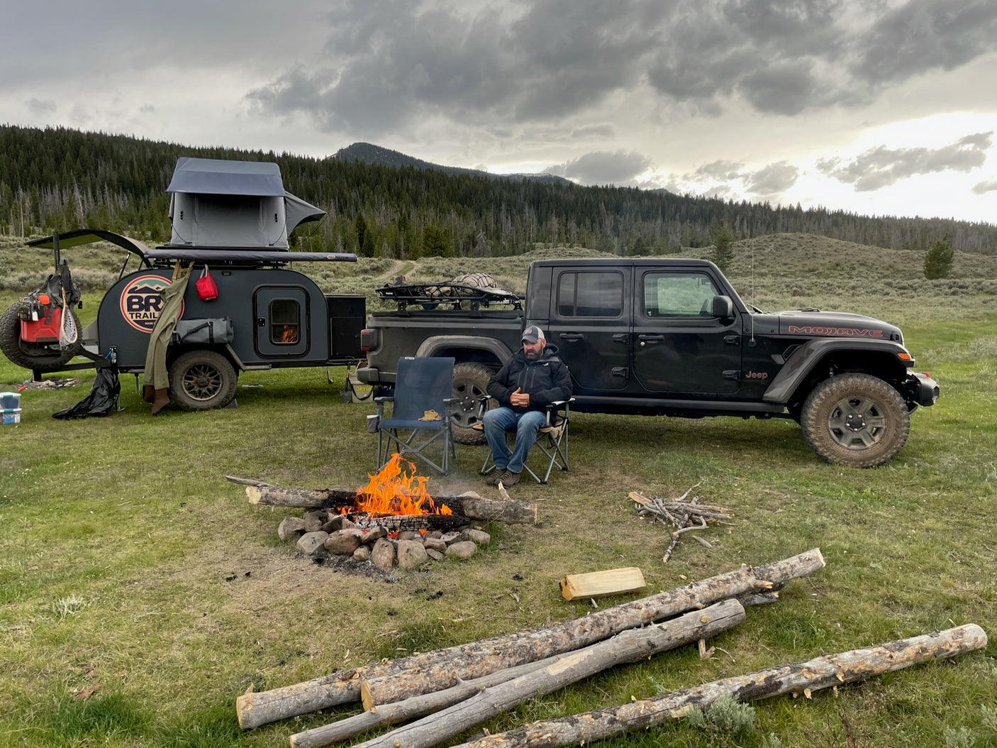 Off-road adventure: Man by campfire with a Jeep & teardrop camper. Explore with an RV rental like this!|This RV rental setup features a black Jeep Gladiator towing a BRX overlanding trailer with a rooftop tent by a scenic mountain lake.|Jeep Gladiator tows a rugged RV rental trailer with a man standing atop