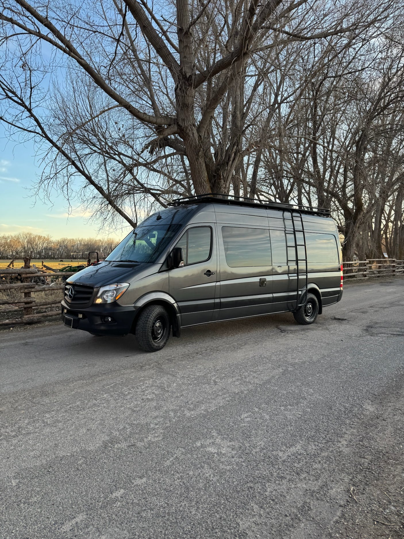 Grey Mercedes Sprinter adventure van on a gravel road, backed by bare trees. Ideal for RV rental getaways!