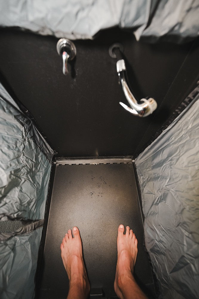 Inside a compact RV rental shower, showing bare feet on the dark floor and chrome fixtures overhead.