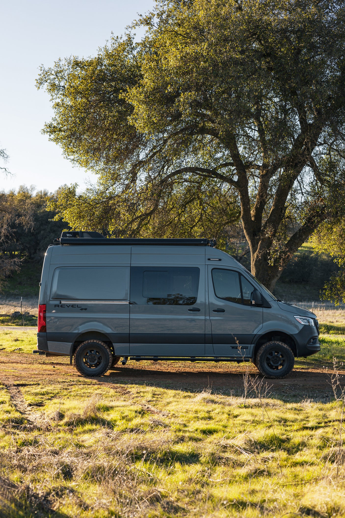 Gray Winnebago Revel 4x4 camper van parked off-road by a tree. Explore nature with this rugged RV rental.