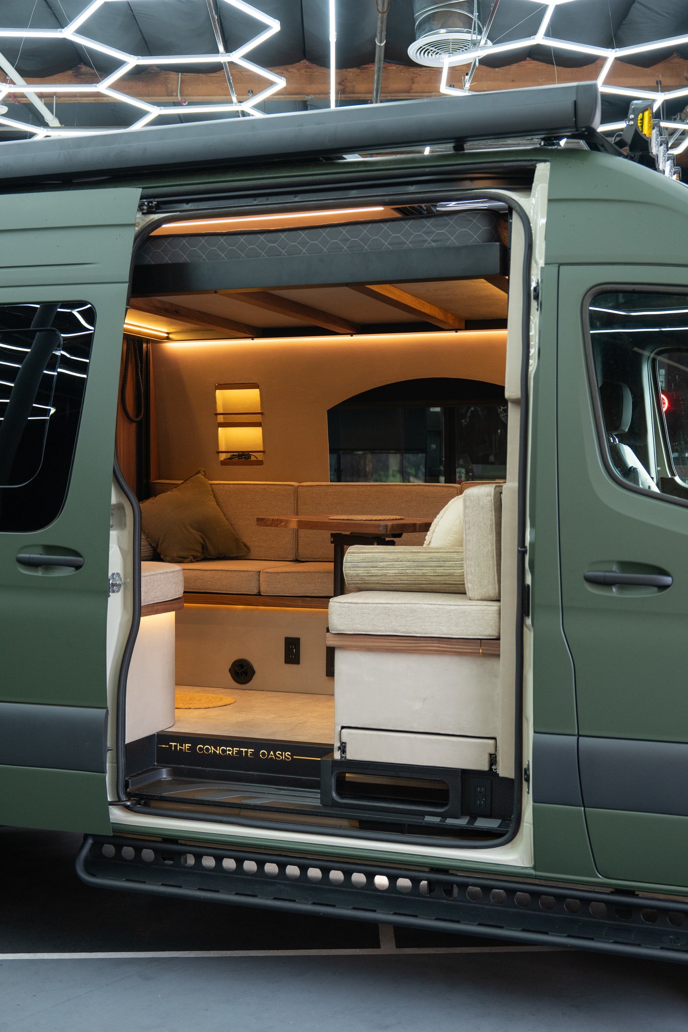 Inside an open RV rental van: a warm, modern custom interior with comfortable seating, table, and overhead sleeping area.
