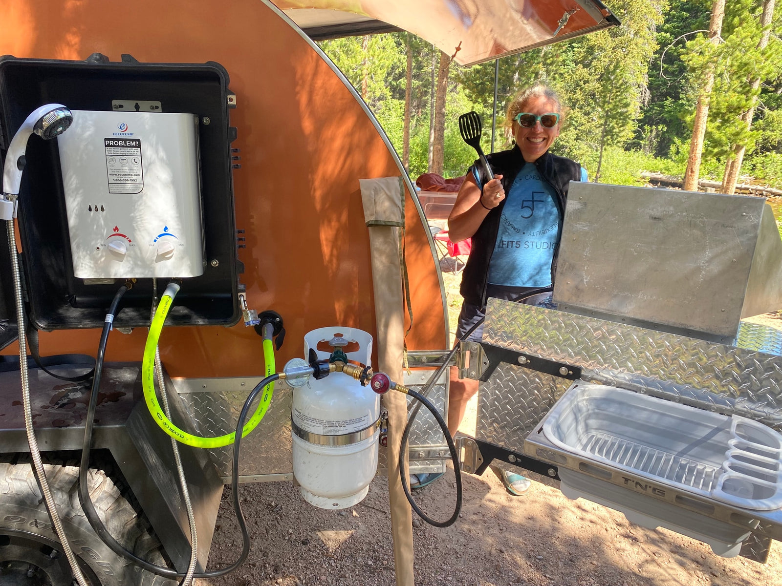 A smiling camper uses a compact outdoor kitchen for an RV rental, featuring a portable water heater & sink setup.