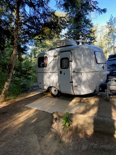A sleek, white travel trailer, ideal for an RV rental, is parked at a scenic campsite among tall trees, with a mat laid out.