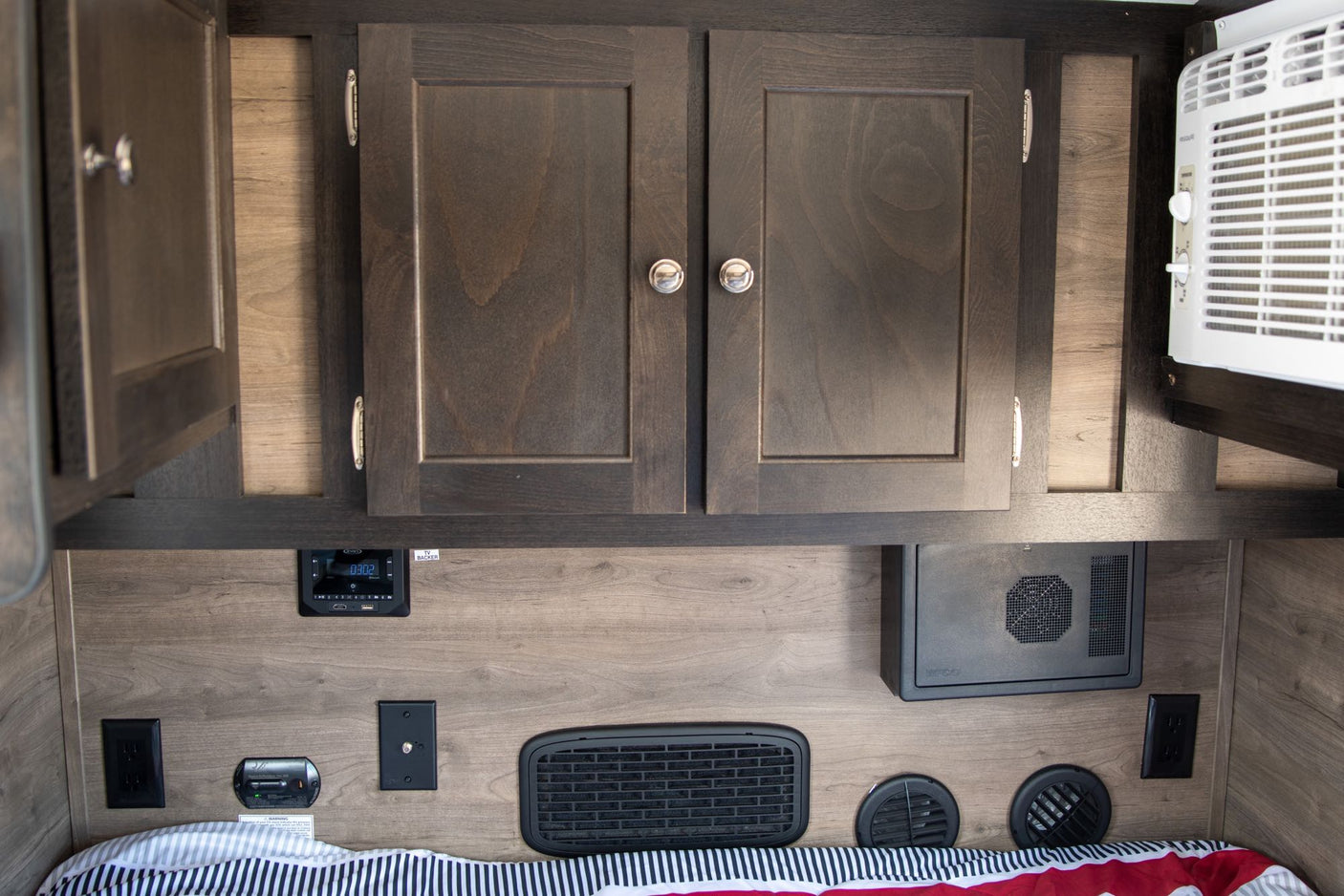 RV rental interior featuring dark wood cabinets, AC unit, control panel, outlets, and vents above a striped bed.