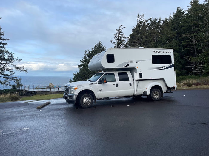 A large white truck camper, ideal for an RV rental, parked on asphalt overlooking the Pacific Coast. Evergreen trees frame the view.
