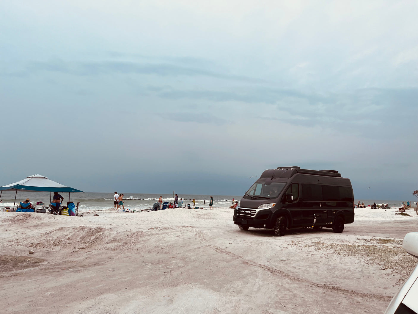 Black RV rental parked on a sandy beach next to the ocean, with people enjoying an overcast day.