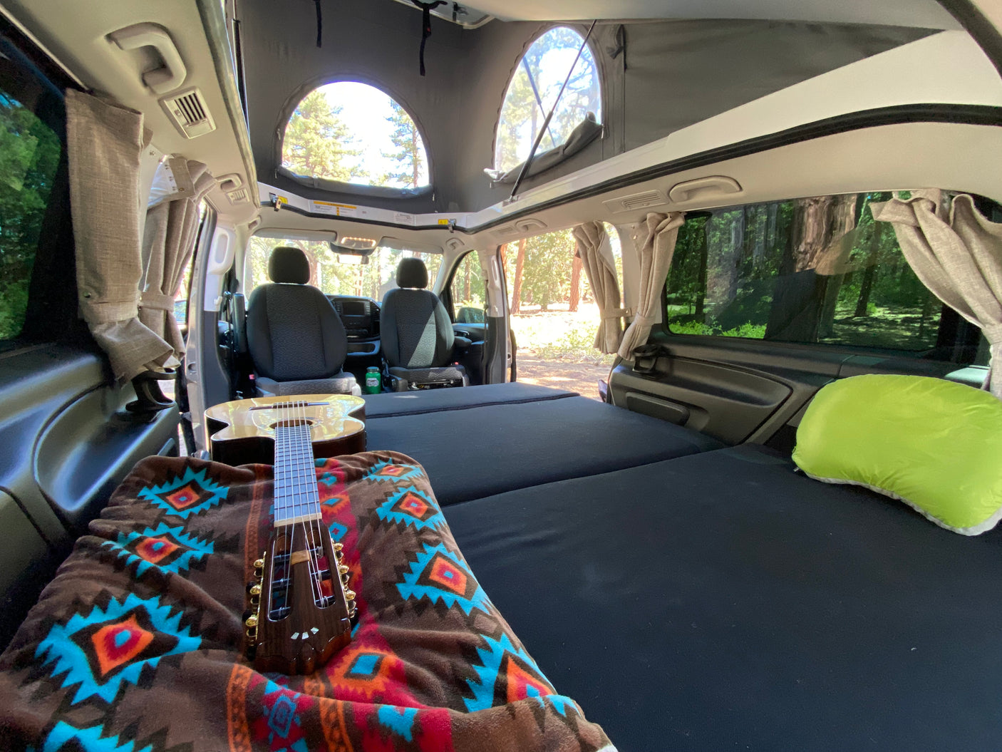Cozy RV rental interior with a pop-top, comfy bed, guitar, and forest views. Ideal for your next camper van adventure!