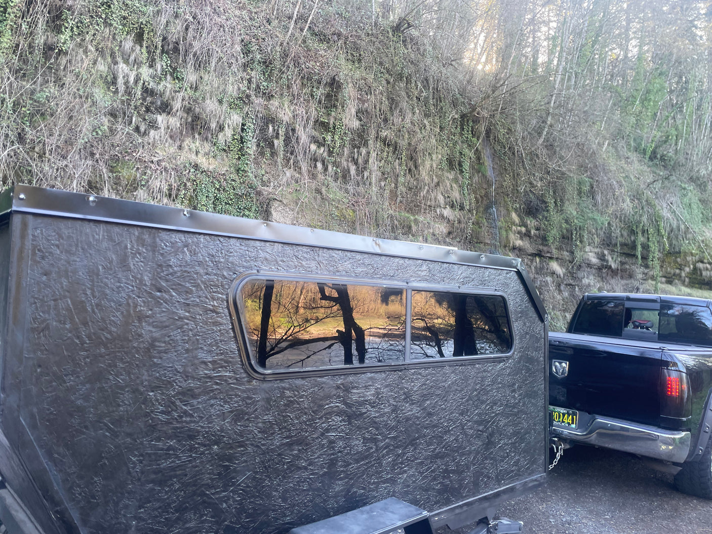 Dark textured teardrop camper/RV rental, towed by a pickup truck, reflects bare trees. Perfect for your next travel adventure.