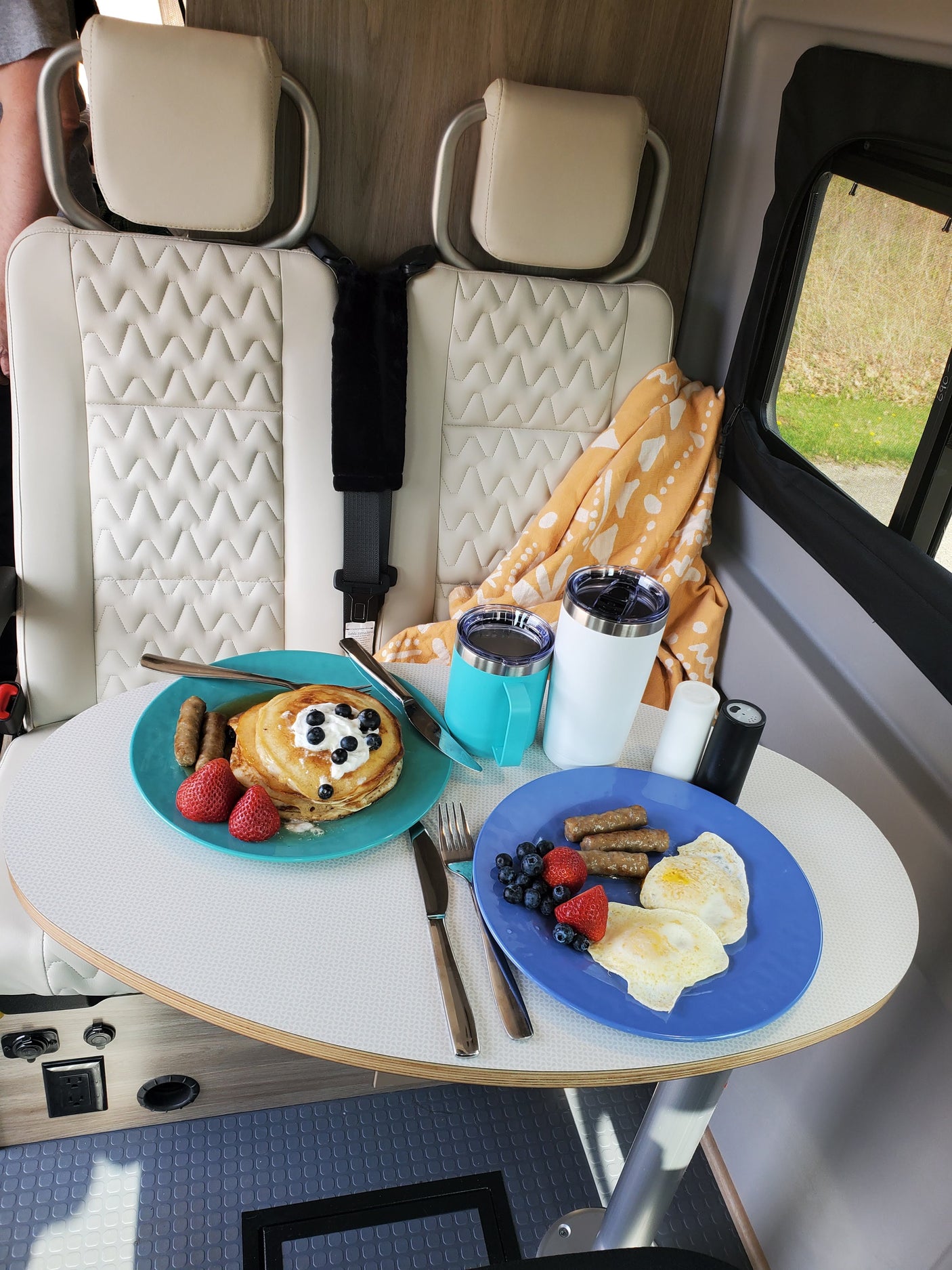 RV rental breakfast scene: two plates with pancakes, eggs, sausage, fruit, and tumblers on a table in a camper van.