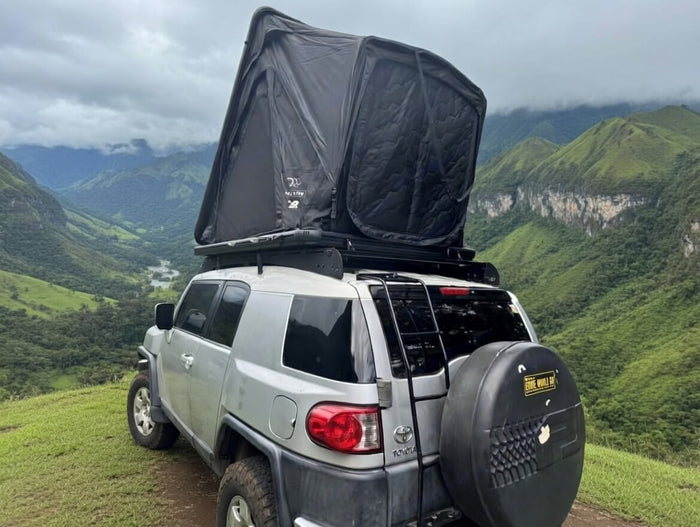 overlooks a stunning green mountain valley under a cloudy sky.|Silver FJ Cruiser with rooftop tent on a stunning beach