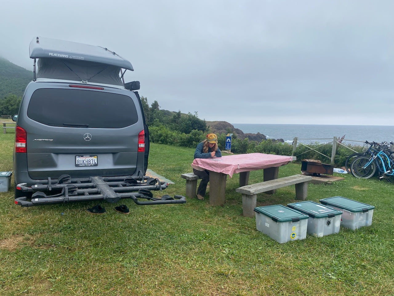 An RV rental camper van parked at an oceanfront campsite. A person sits at a picnic table; bikes are nearby.