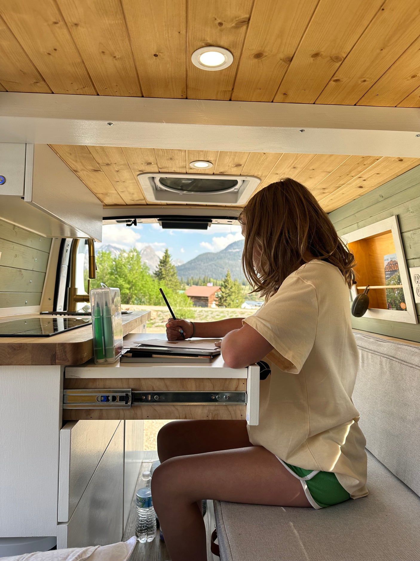 Cozy RV rental workspace: girl drawing at a desk with mountain views. Ideal for van life travel & creativity.