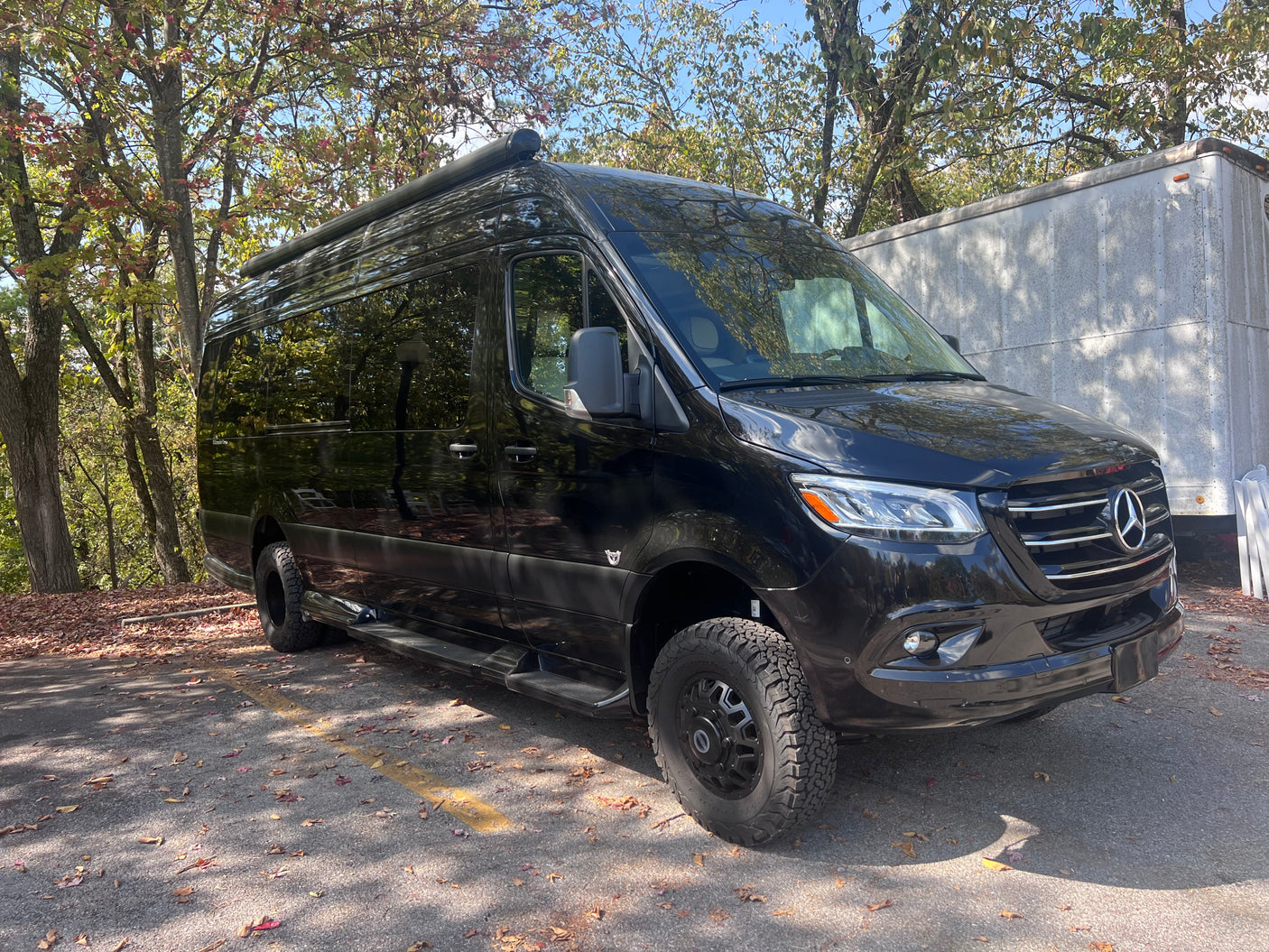 Black Mercedes-Benz Sprinter van RV rental, equipped with an awning & off-road tires, parked amidst autumn trees.