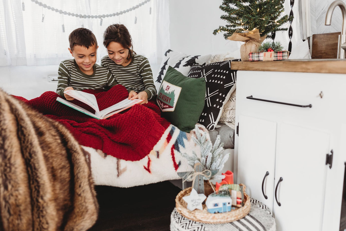 Cozy RV rental scene: Two kids reading a book under a red blanket, surrounded by holiday decor in their tiny home.