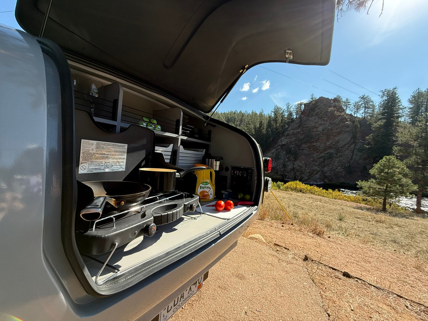 RV rental kitchen setup with stove, orange juice, and tomatoes, overlooking a scenic river and mountains.