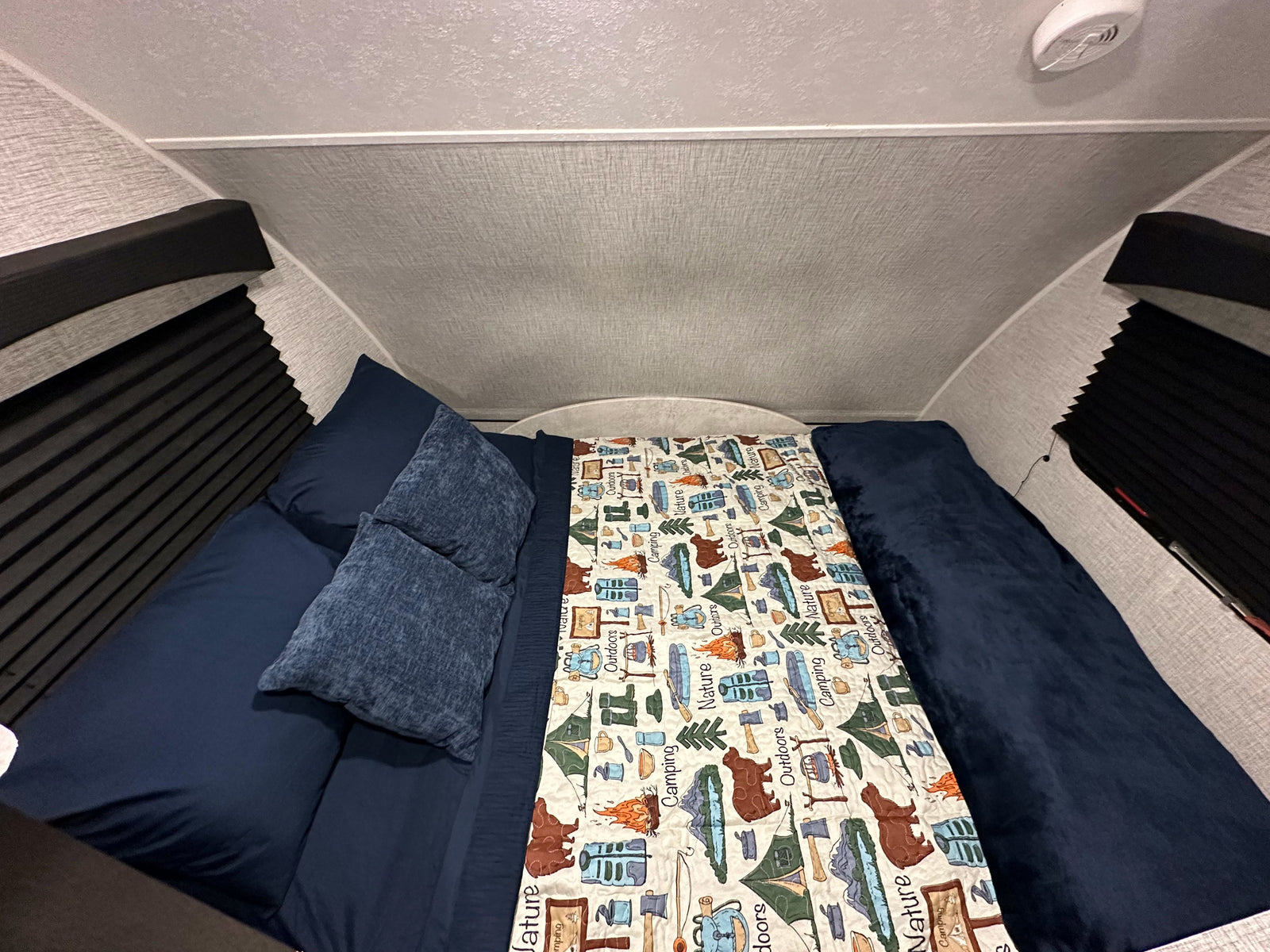 Cozy RV rental bedroom with dark blue bedding and a nature-themed quilt, featuring compact design and window blinds.