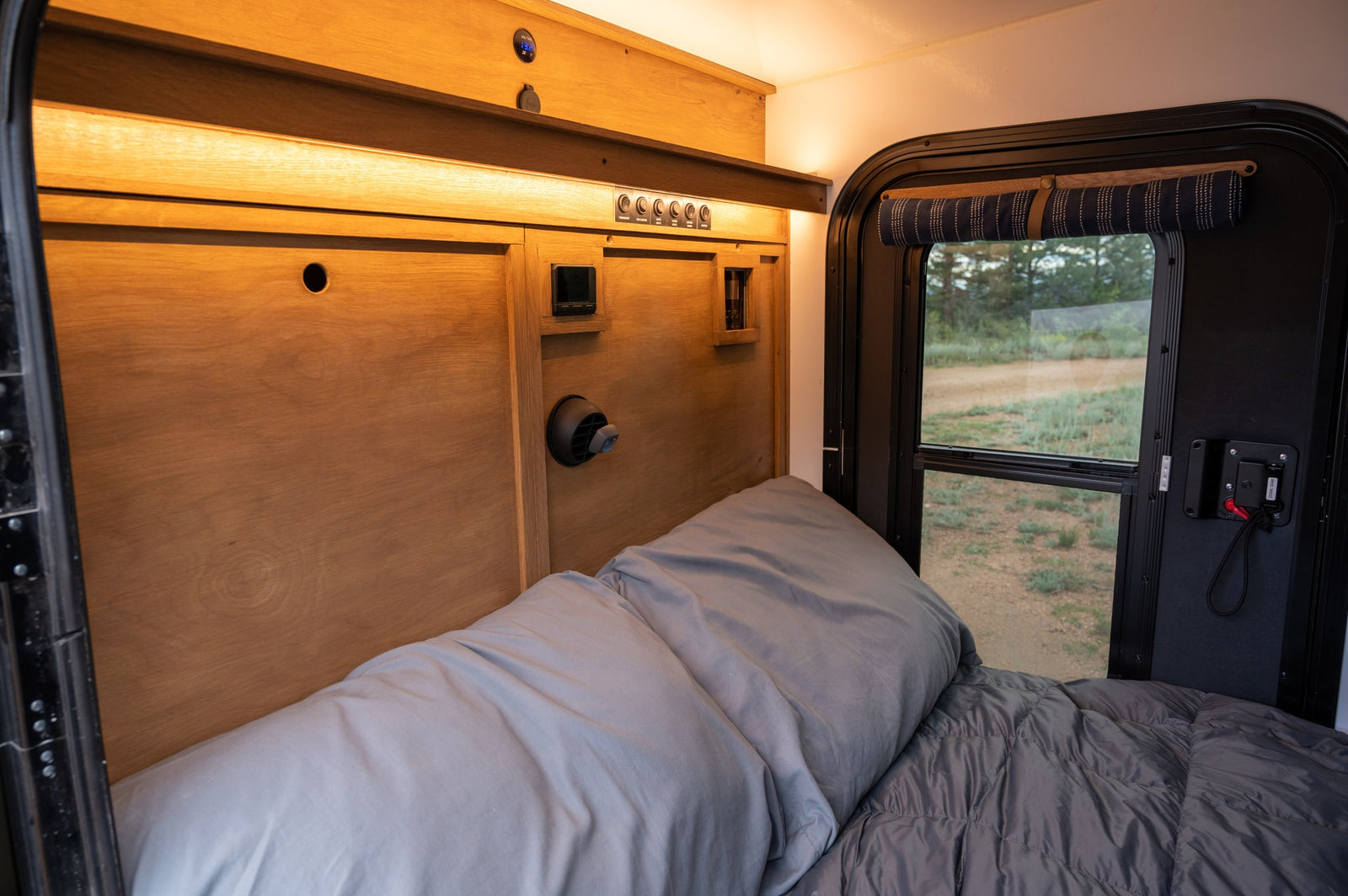 This RV rental offers a cozy bed with light grey bedding, warm wooden walls, essential controls, and a window view.