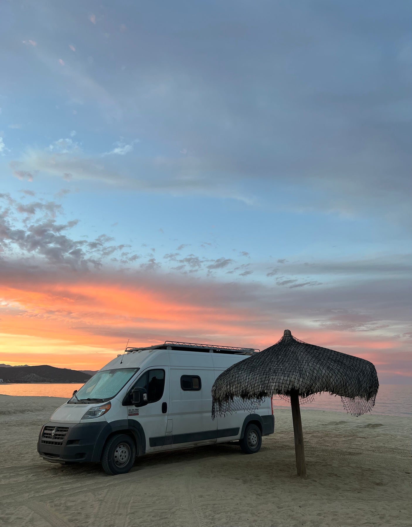 Stunning beach sunset with an RV rental. White camper van parked on sand next to a palapa by the ocean.