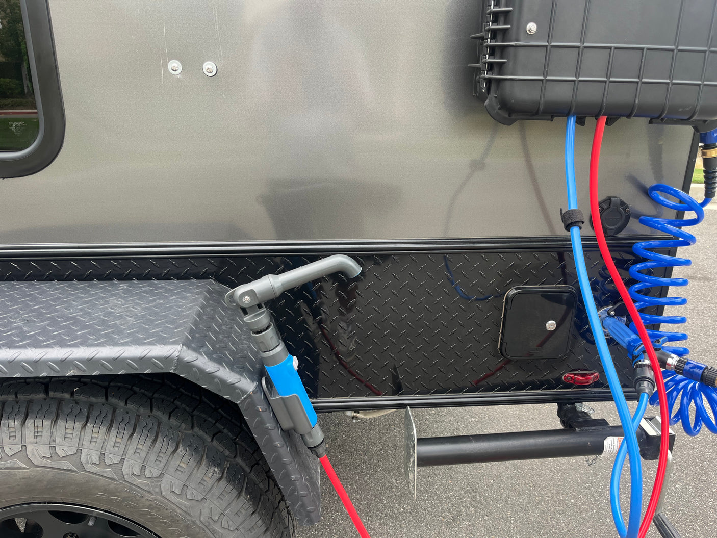 RV rental: Outdoor shower system with blue/red hoses, coiled hose, storage box, and tire on a grey/black trailer.