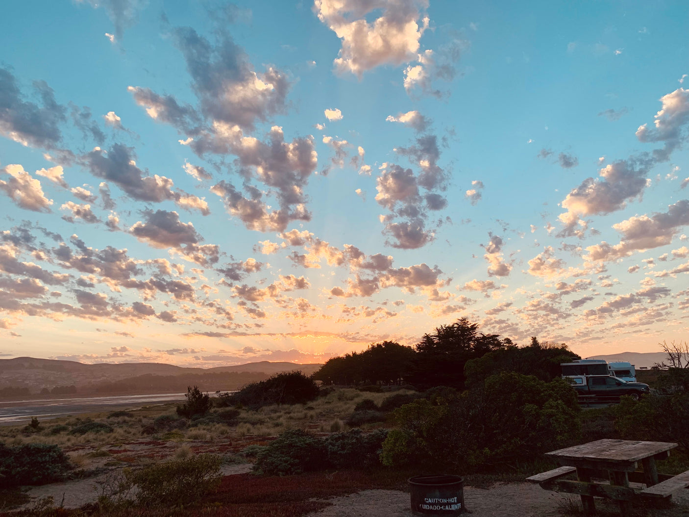 Golden hour at a tranquil campsite, showcasing an RV rental, picnic table, and fire pit by the bay.