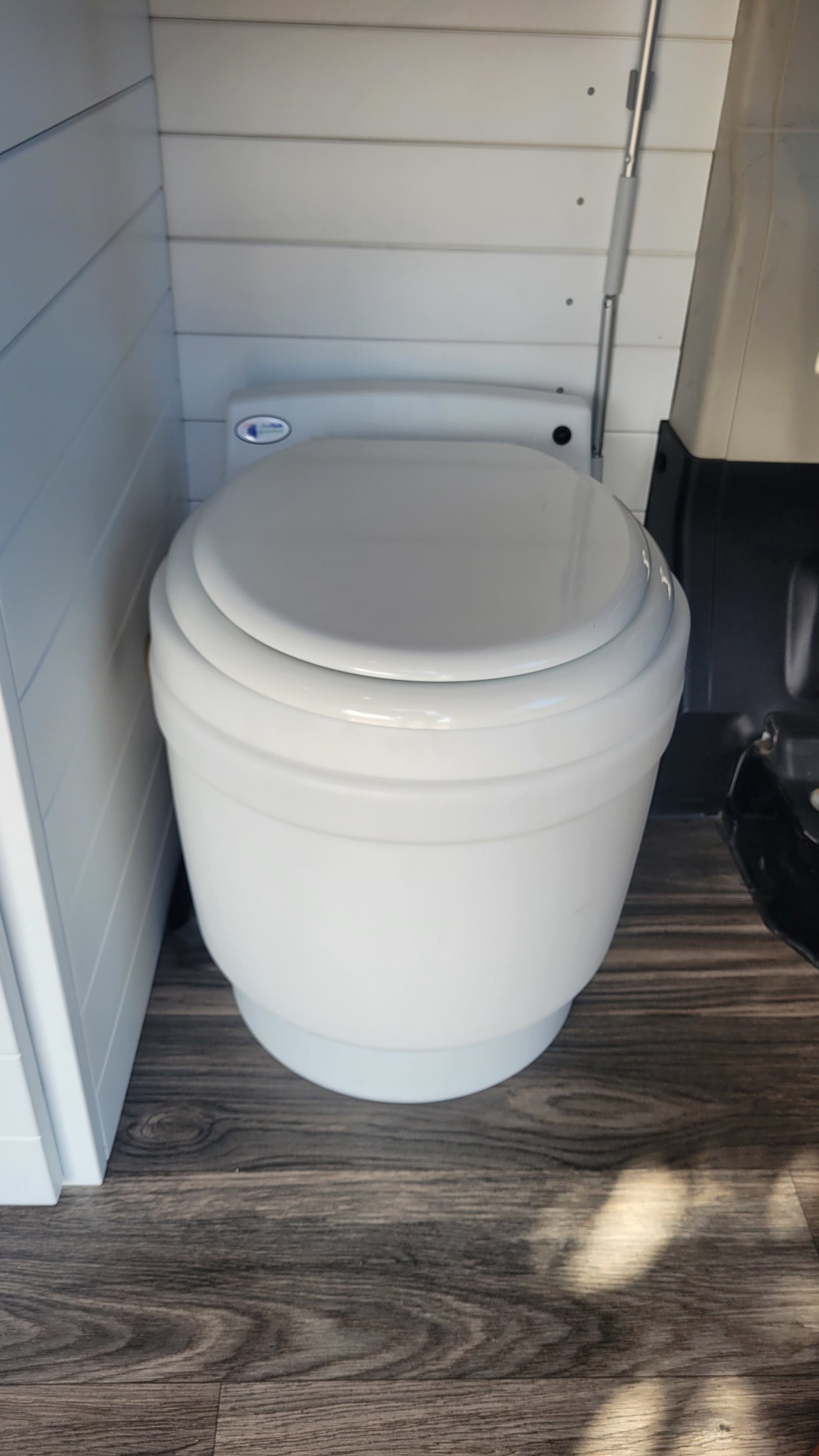 White portable toilet with closed lid on wood floor, installed in an rv rental vehicle with white slatted walls.