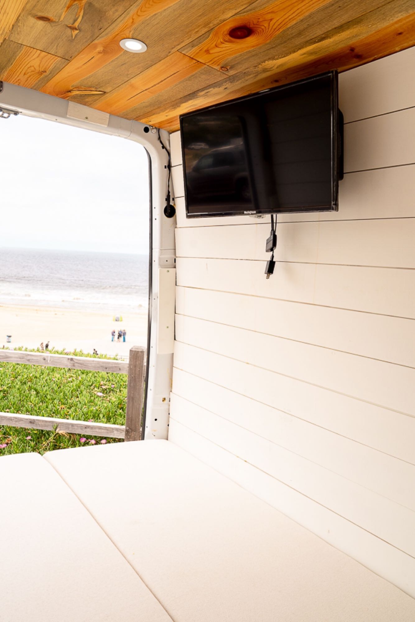 RV rental interior with a view of the beach, featuring a mounted TV, wooden ceiling, and comfy bed.