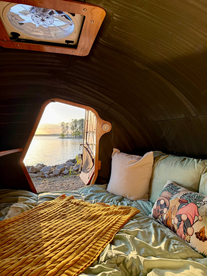 a comfy bed faces an open door with a beautiful lake & sunset view. Ideal for camping.|Cozy RV rental interior looking out at a sparkling lake and cloudy sky. Person's hands clasped with smartwatch.|**RV rental** lake view from inside a camper: open door reveals a sandy campsite with rocks
