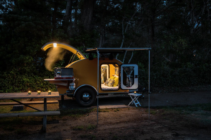 An illuminated yellow teardrop RV rental camper at night, featuring a cozy interior, open cooking area, and picnic setup.