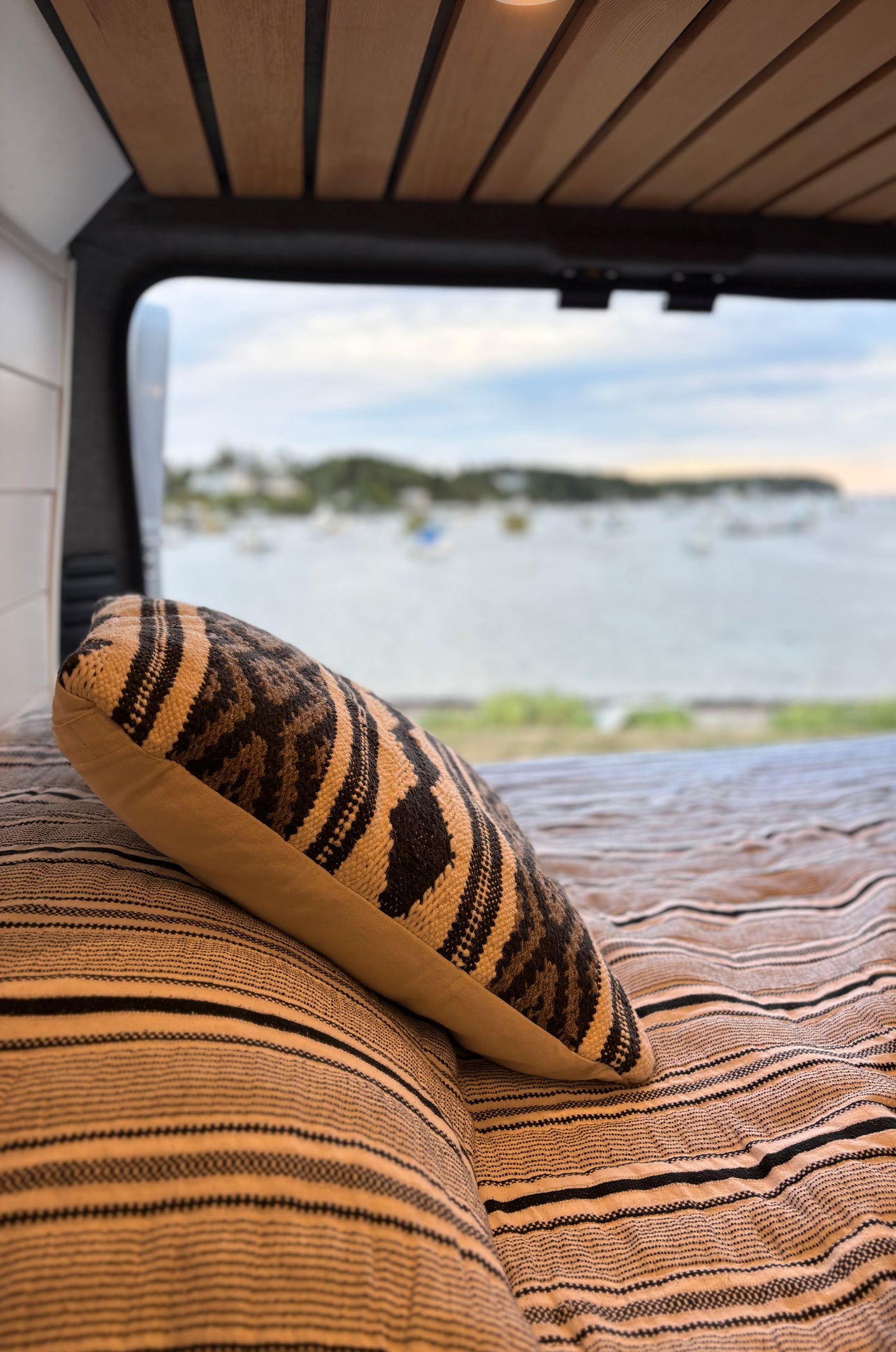 Cozy RV rental bed with a patterned pillow and a view of a tranquil bay with anchored boats.