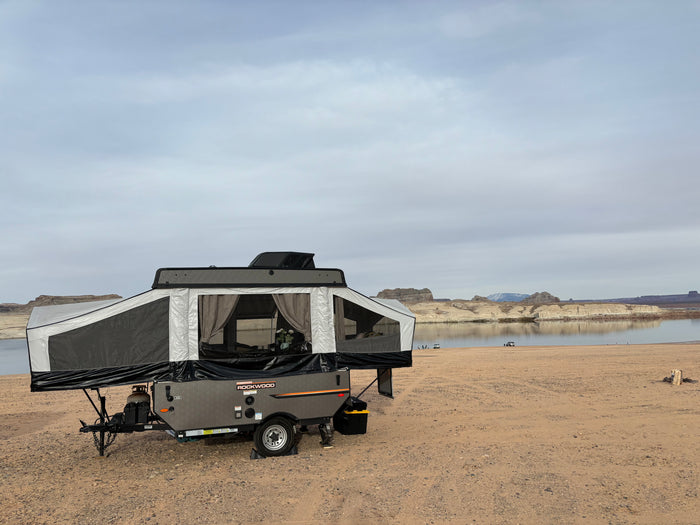 An RV rental, a grey Rockwood pop-up camper, is set up on a sandy beach next to a calm lake with cliffs beyond.
