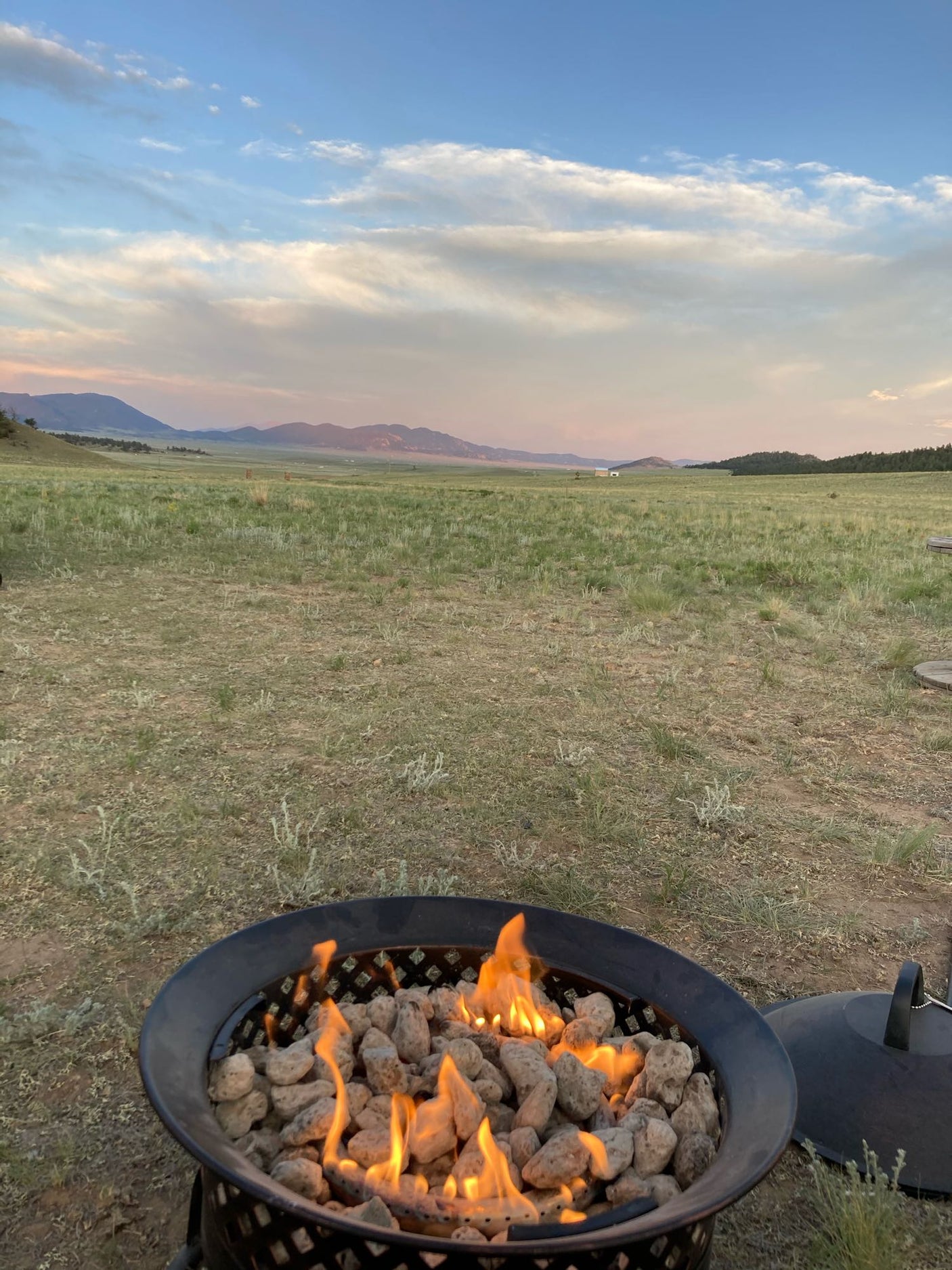 RV rental perfect for a peaceful evening: a warm fire pit burning, vast fields, and distant mountains under a pastel sky.