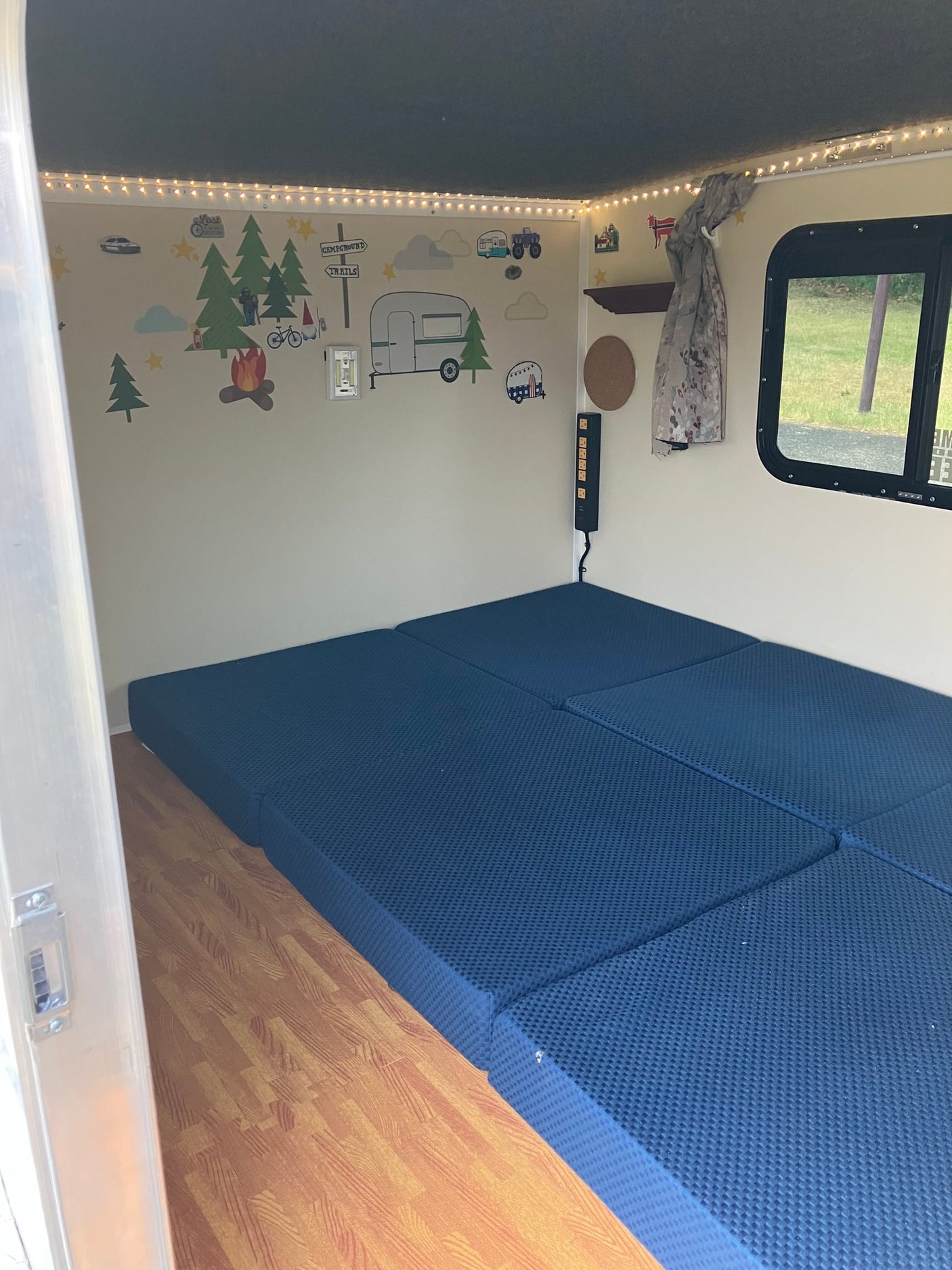 Inside an RV rental: decorated walls, comfy blue modular bed, string lights, and a window view. Perfect for camping trips.